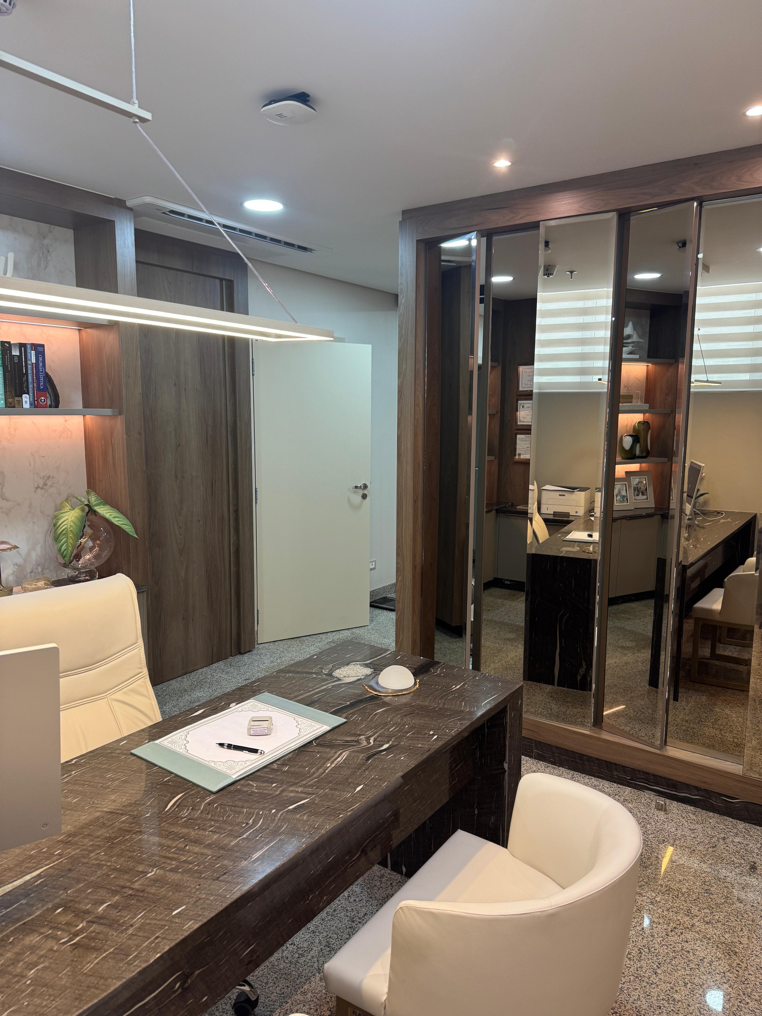 Sala de consulta vista de outro ângulo, com mesa de mármore, poltronas claras, luminária pendente e parede espelhada